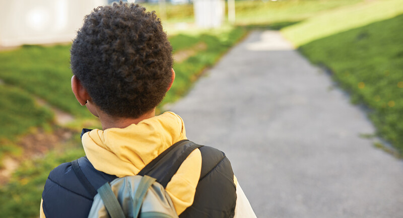Photo from behind of a young boy walking on a sidewalk, wearing a backpack