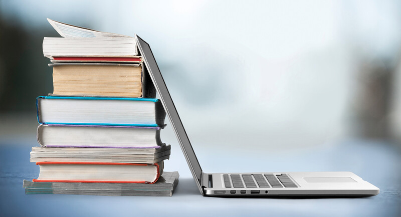 Photo of a laptop sitting open with its screen leaning back against a stack of books.